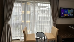 Habitación de hotel con un ventanal grande, dos sillones color beige, una mesita redonda y un televisor de pantalla plana montado en la pared.