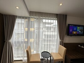 A hotel room with a large window, two beige armchairs, a small round table, and a flat-screen TV mounted on the wall.