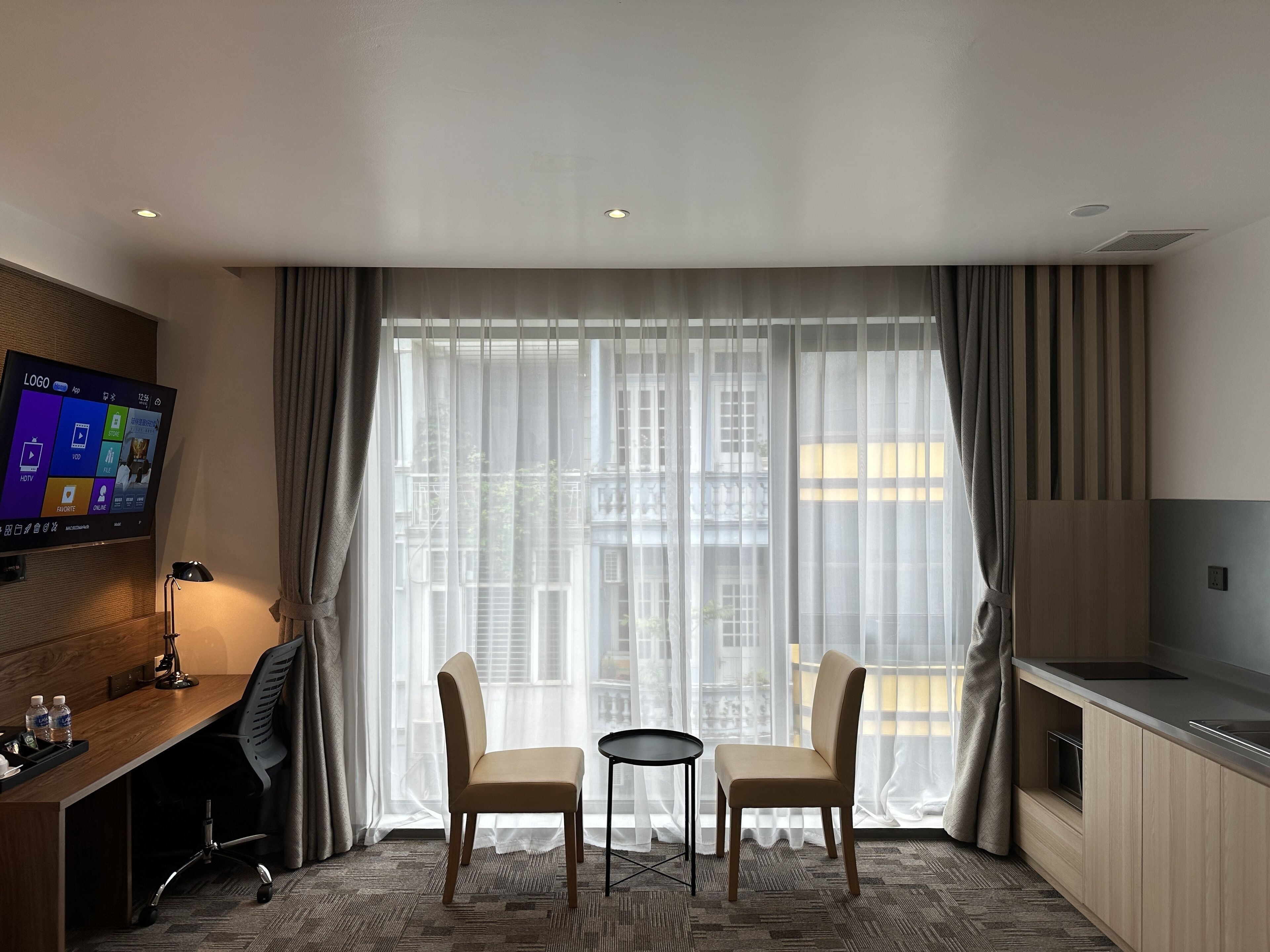 A hotel room with a large window, a desk with a chair, a TV, and a kitchenette.