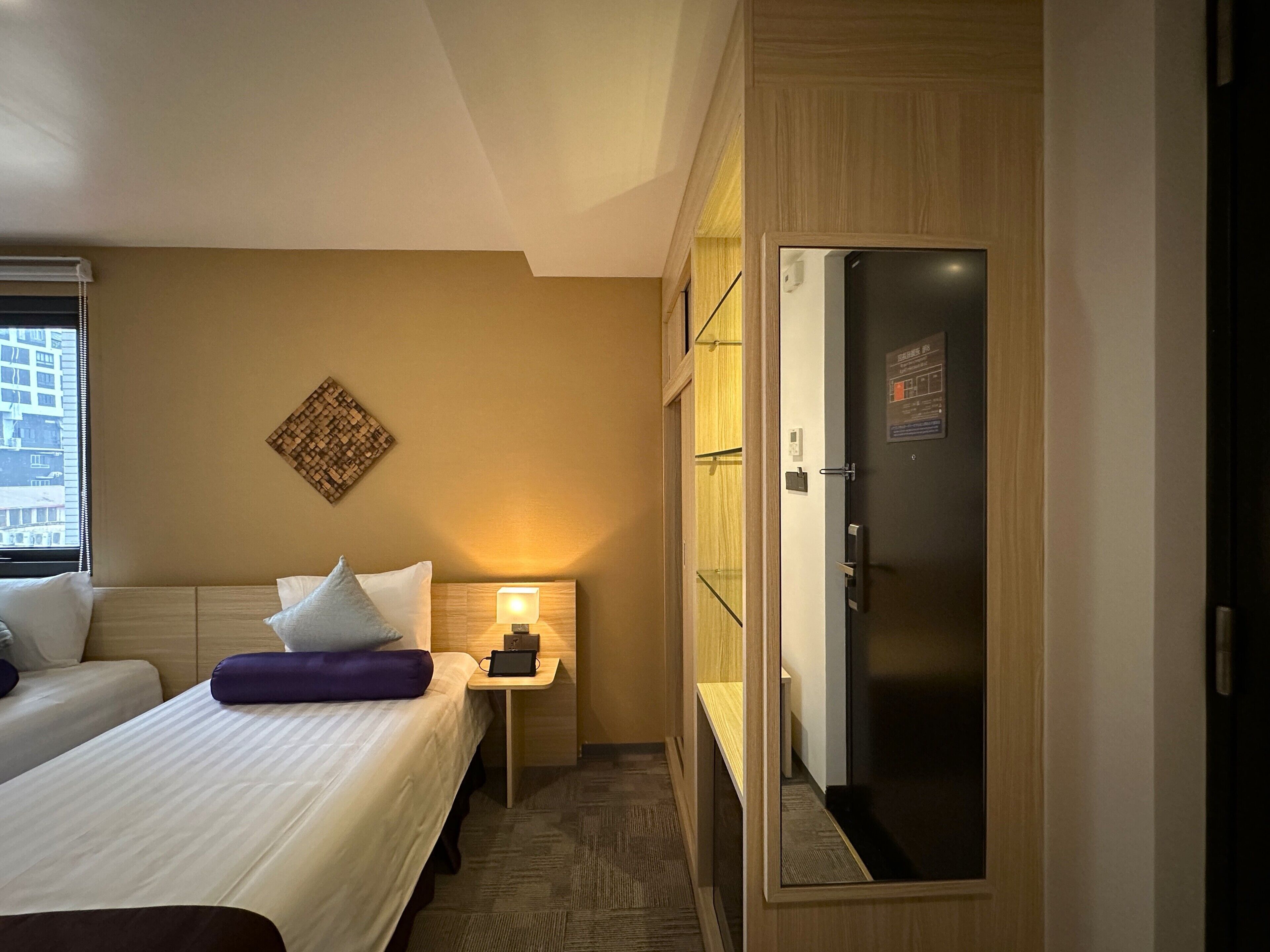 A hotel room with two beds, a nightstand with a lamp, a minibar, and a view of the city through the window.