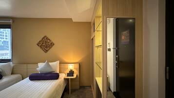 Habitación de hotel con dos camas, una mesita de noche con lámpara, minibar y vista a la ciudad desde la ventana.