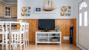 A modern kitchen with white cabinetry, a dining table with chairs, a flat-screen TV on a wooden stand, and nautical-themed artwork.