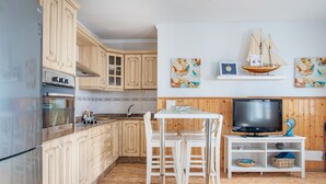 A kitchen with wooden cabinets, a built-in oven, a dining table with chairs, a TV on a wooden stand, and artwork on the wall.