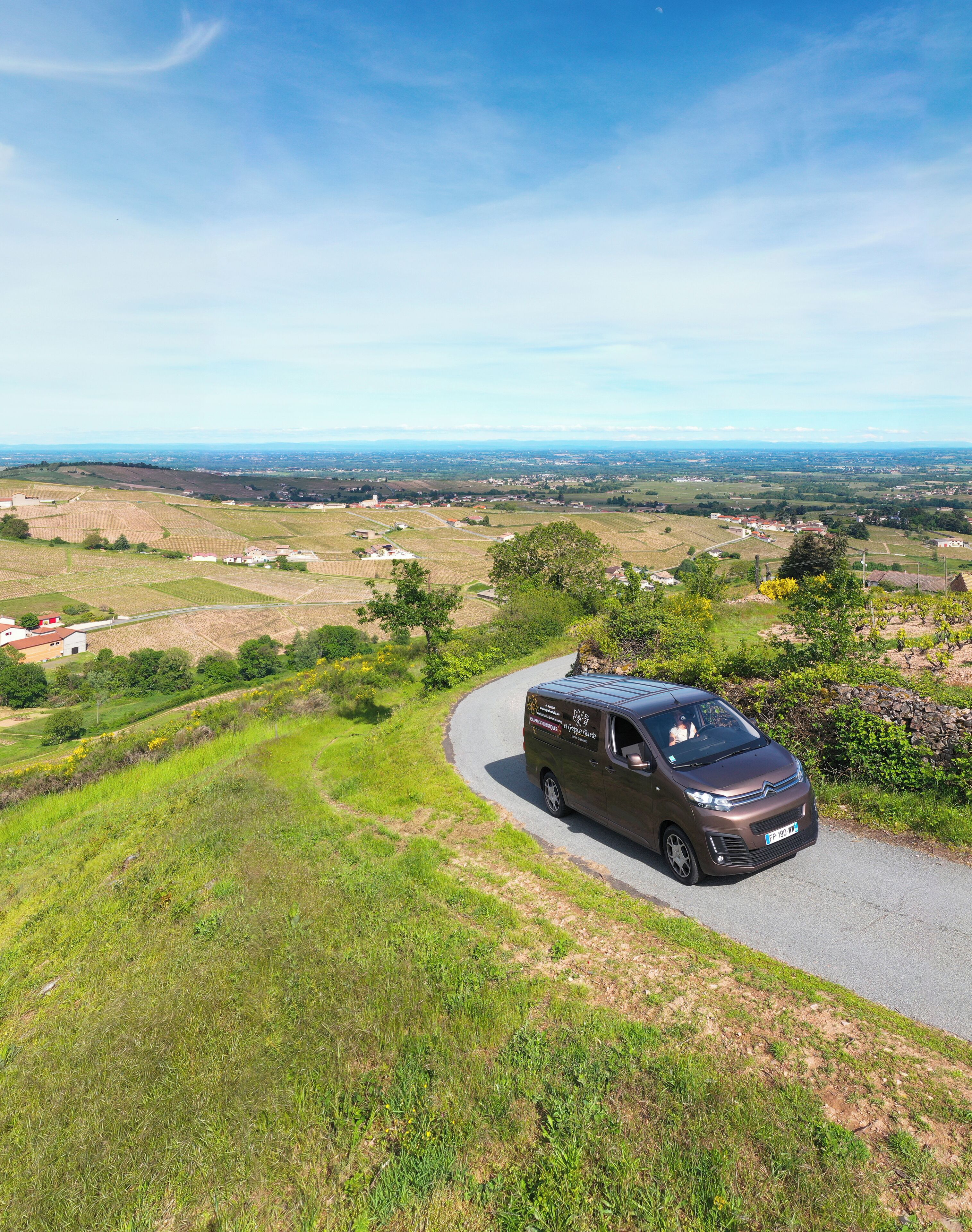 A brown van parked on a winding road with a view of a rural landscape, fields, and distant buildings.