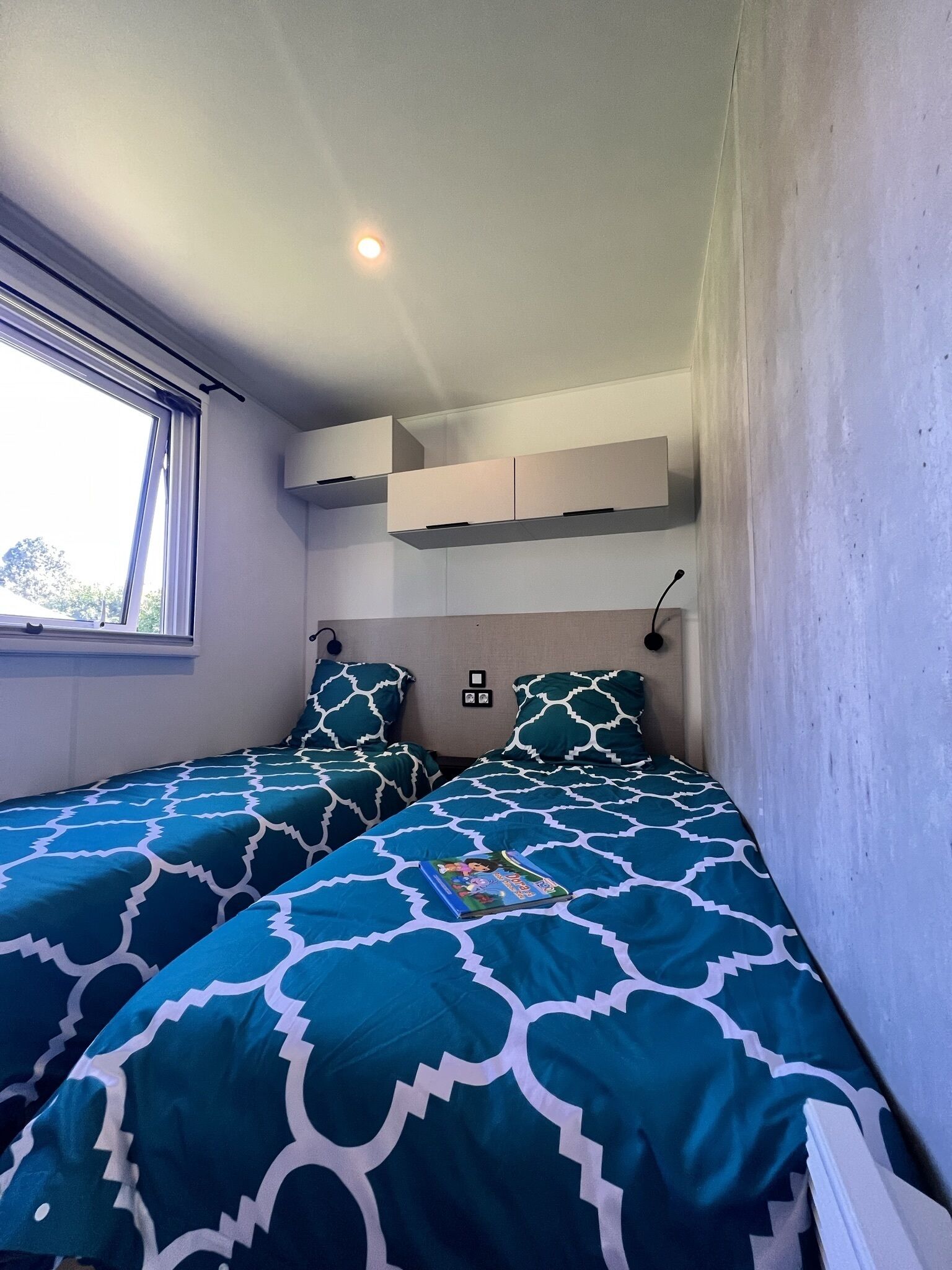 A small, well-lit bedroom with two beds, a window, and built-in storage.