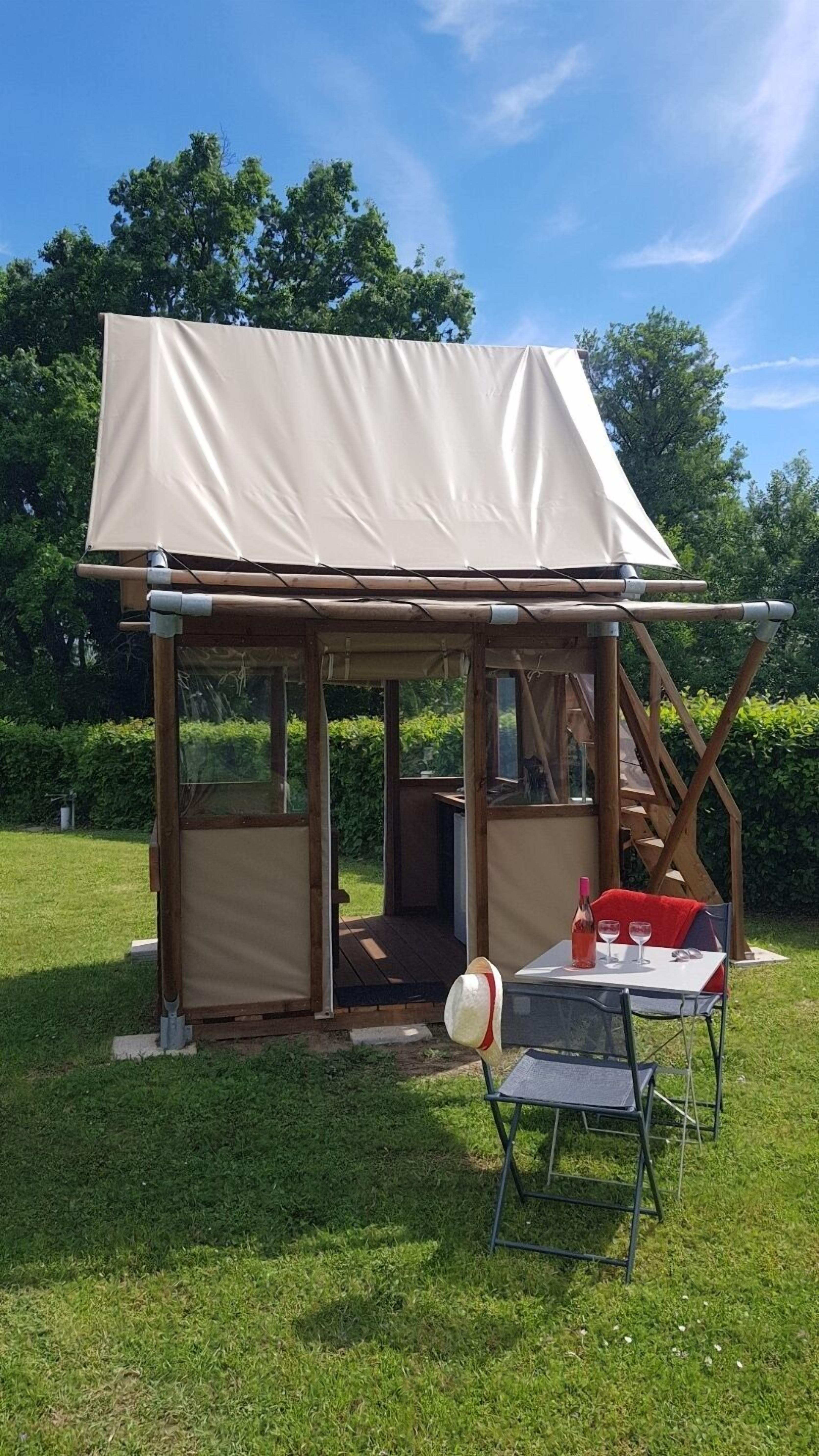 A small wooden structure with a canvas roof, a table with chairs, and a bottle of red wine.