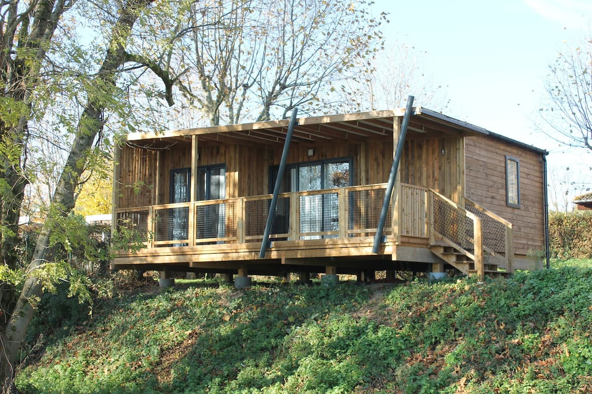 A wooden cabin with a balcony, situated on a hillside with trees around.