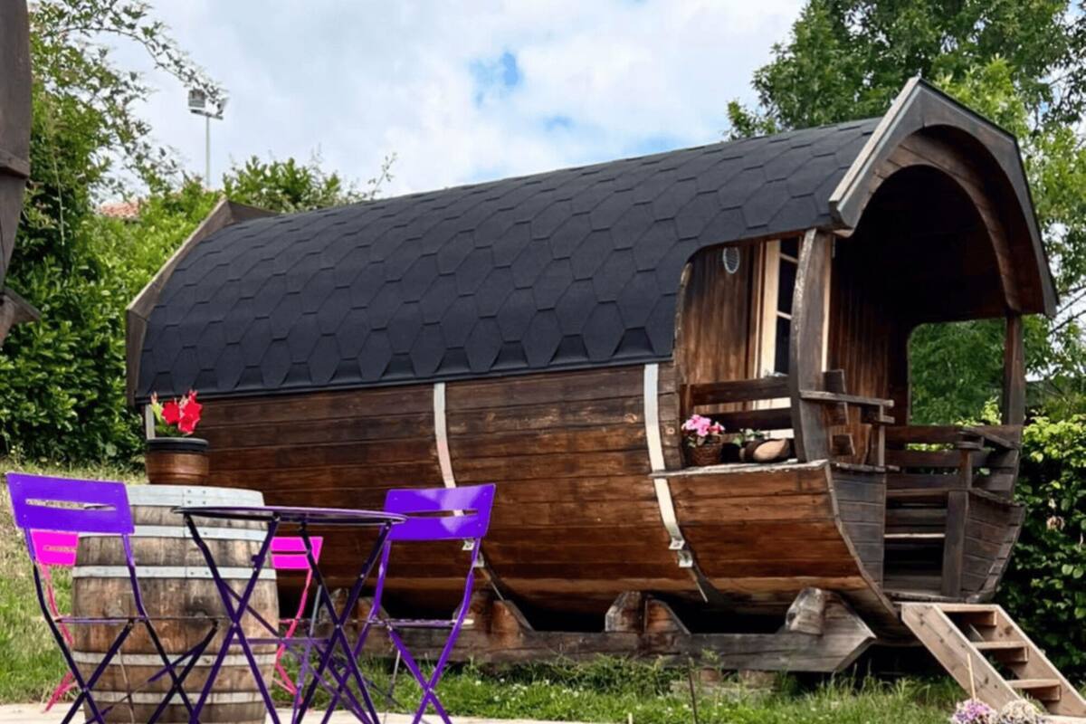 A wooden structure with a sloped roof, possibly a cabin or a small house, with a staircase leading up to it. There are two purple chairs and a small table in front.