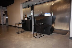 A modern hotel reception area with a black reception desk, a digital information kiosk, and a seating area with a small table and chairs.