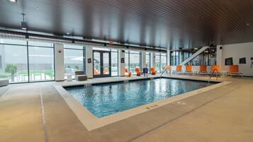 Una piscina interior con agua cristalina, rodeada de sillones de descanso naranja y blanco, y vistas al exterior a través de amplias ventanas.