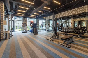 Un gimnasio con varios equipos de ejercicio, ventanas amplias y una alfombra estampada.