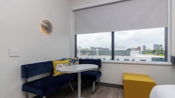 Une pièce moderne avec un canapé bleu, une petite table ronde et un pouf jaune. Une fenêtre offre une vue sur le paysage urbain.