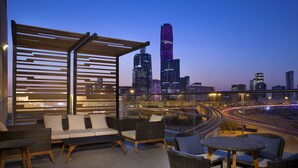 Terraza en la azotea con mobiliario de mimbre, barandilla de vidrio y vistas a la ciudad al anochecer.
