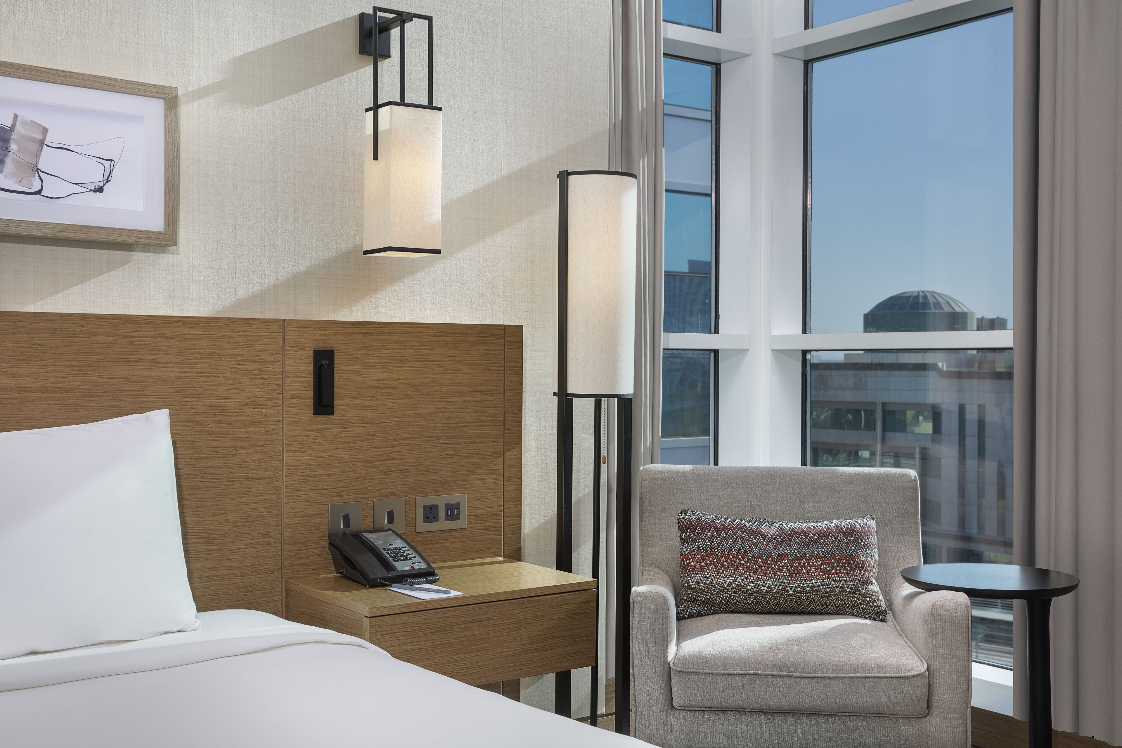 A hotel room with a bed, a desk with a phone, a chair with a patterned pillow, a standing lamp, and a large window with a city view.