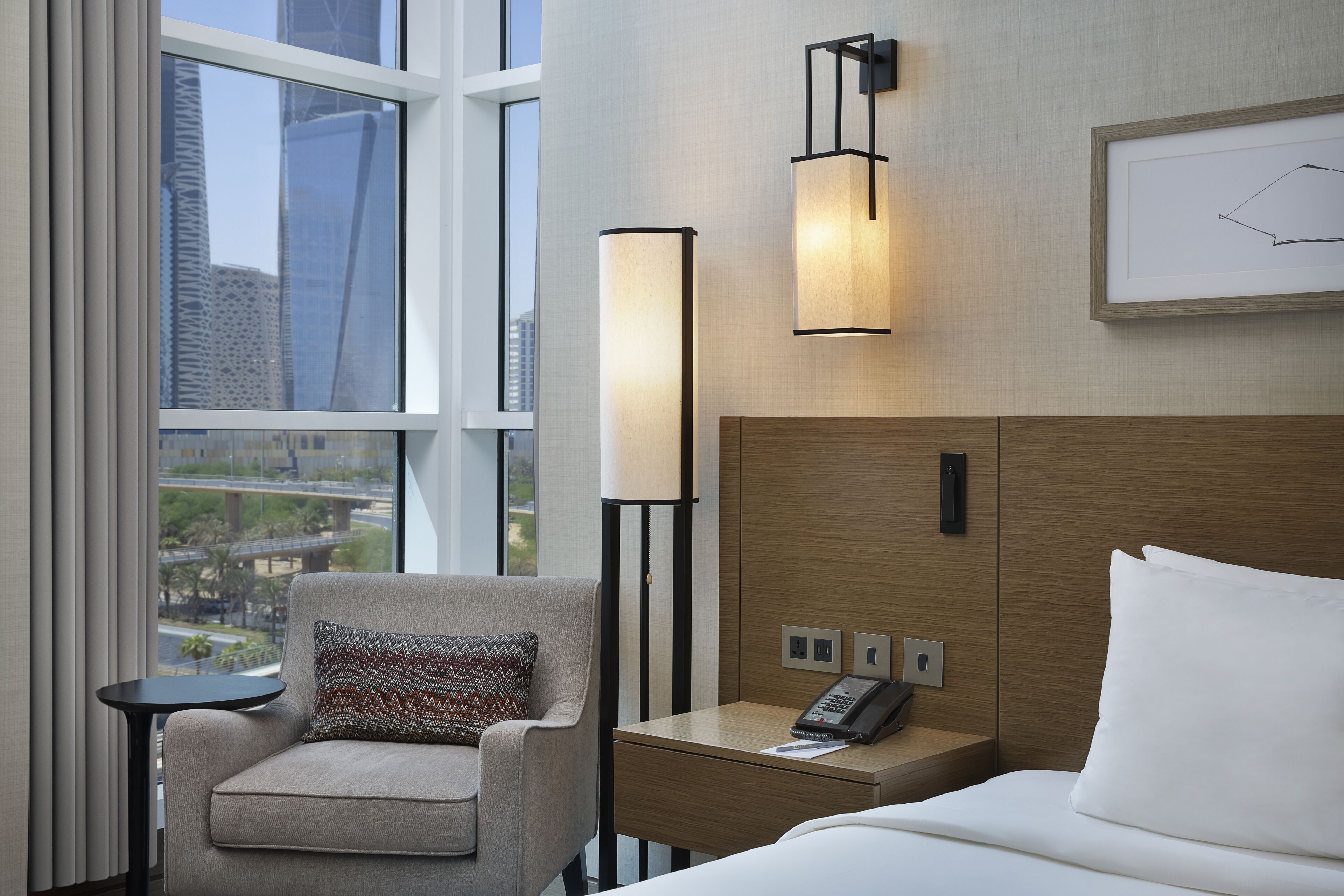 A hotel room with a bed, a chair, a bedside table with a phone, a lamp, and a window with a view of buildings.