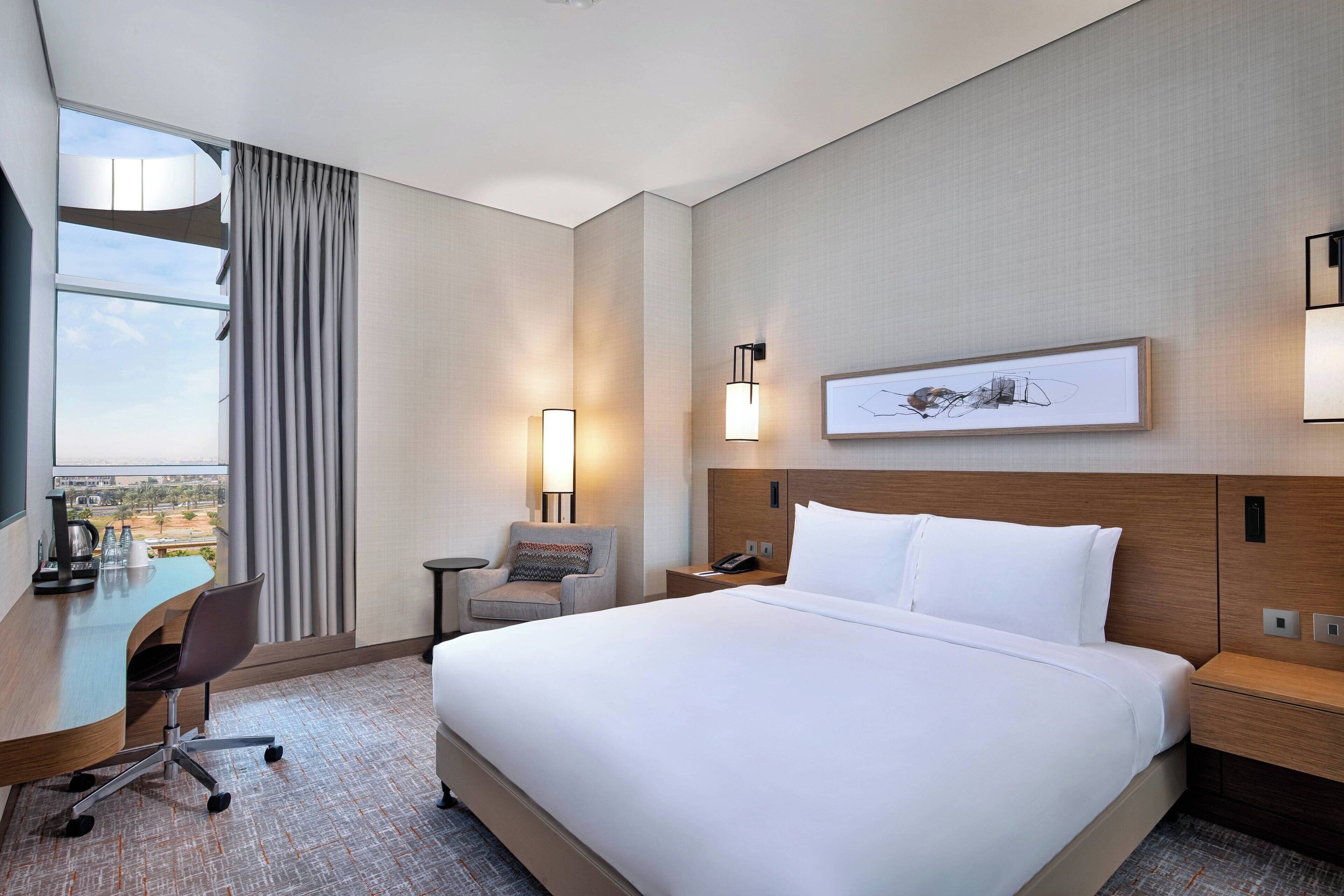 A hotel room with a large bed, a desk with a chair, a small side table, a nightstand, a lamp, and a framed picture on the wall.