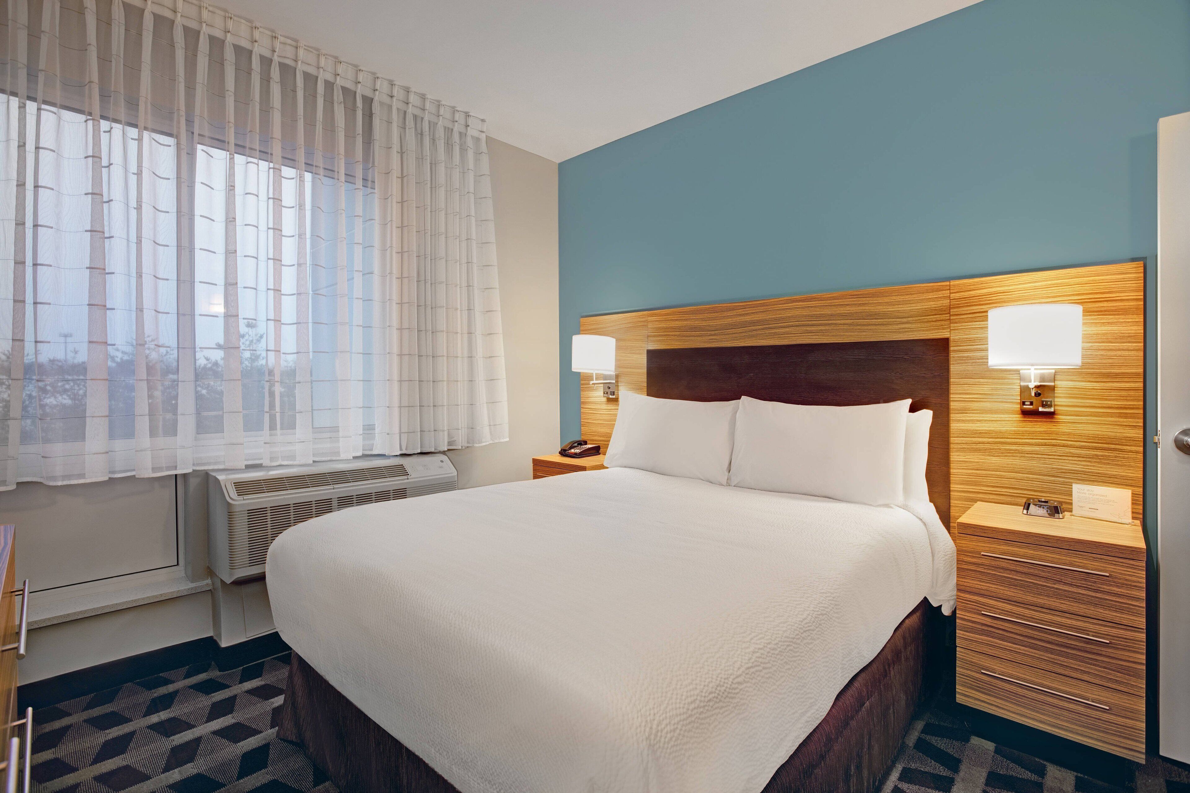 A hotel room with a large bed, two bedside lamps, a wooden headboard, and a window with sheer curtains.