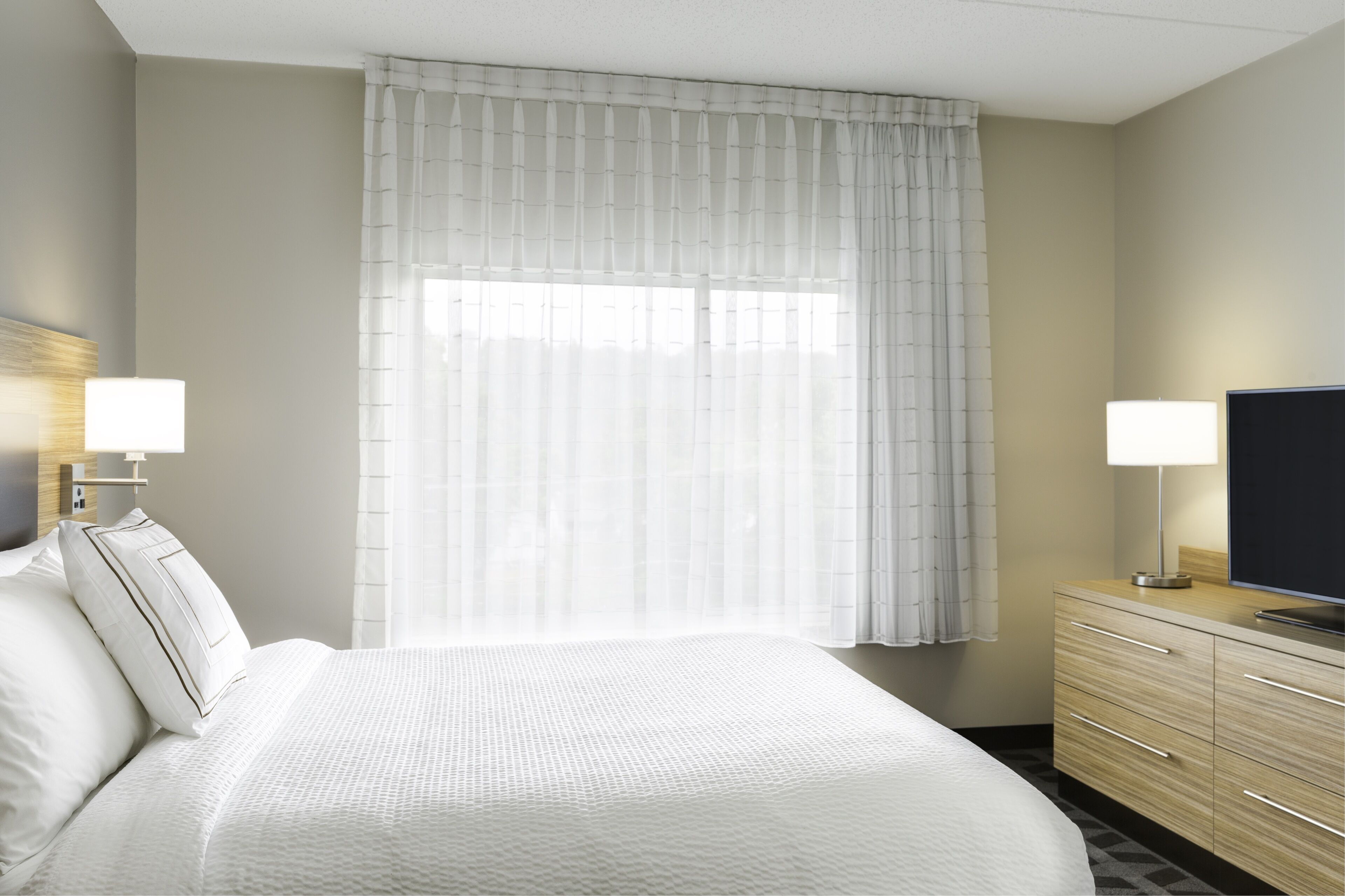 Una habitación de hotel con una cama grande, dos lámparas de noche, un televisor de pantalla plana, un aparador de madera y cortinas translúcidas.