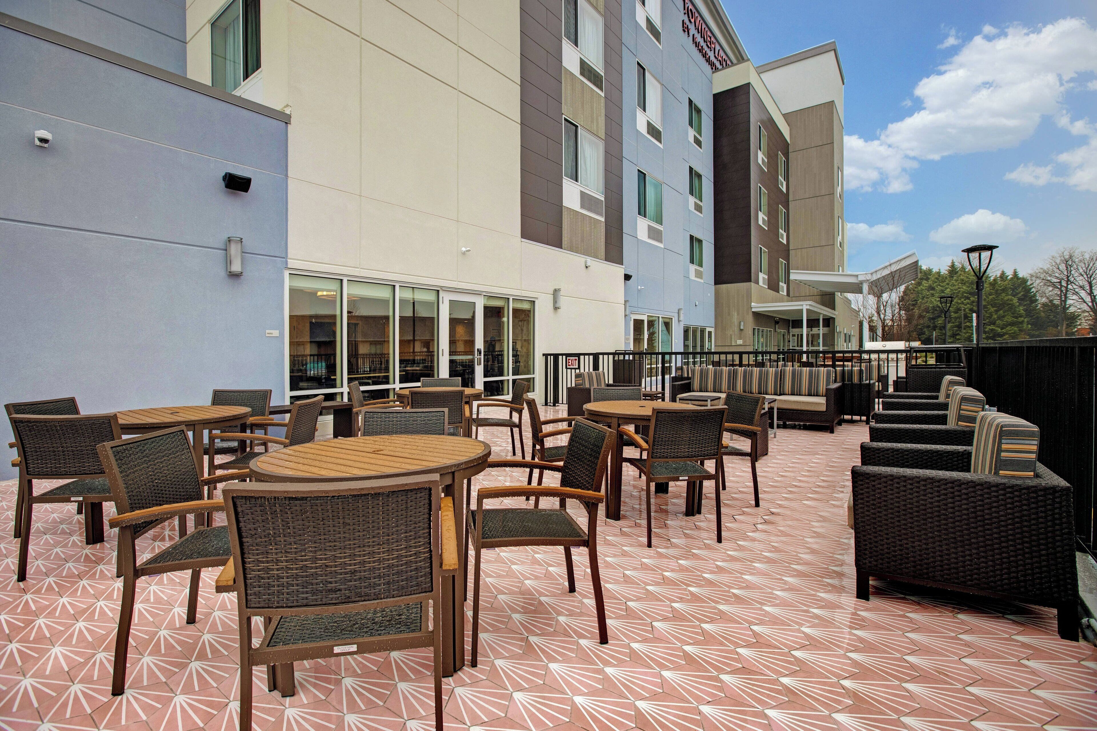 Un espace extérieur aménagé en terrasse, avec des tables et des chaises rondes, un coin salon abrité et un bâtiment doté de nombreuses fenêtres et d’une enseigne.