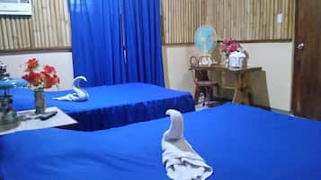 A room with two beds covered in blue sheets, a wooden desk with a fan, a chair, and a small table with a vase of flowers.