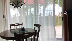 A dining area with a round table, four chairs, a chandelier, and a view through sheer curtains to an outdoor deck.