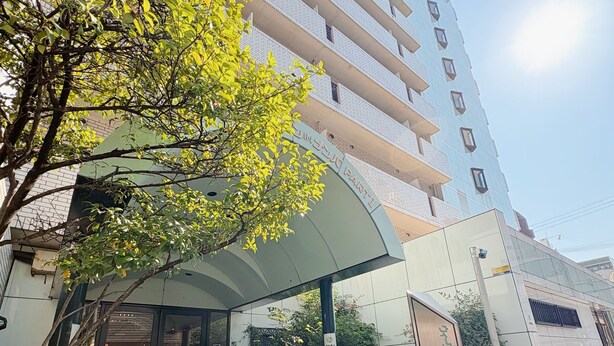 A modern building with a covered entrance, a green carpeted area, and a tree in the foreground. - Weekly Green In Namba (Osaka)
