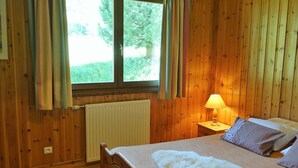 A wooden-paneled room with a bed, a window with curtains, a radiator, and a table with a lamp. - Ski-in/Ski-out Apartment, 2-Bedroom, Sleeps 6 with South Balcony and Parking (Arêches-Beaufort)