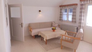 Ein kleiner, gut beleuchteter Raum mit einer Couch, einem Tisch mit Blumenvase, einem Sessel und einem Fenster mit Vorhängen.
