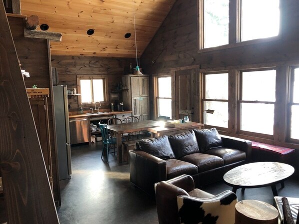 A rustic cabin interior with a wooden ceiling, a dining area with a table and chairs, a leather sofa, and a kitchen with cabinets and a refrigerator.
