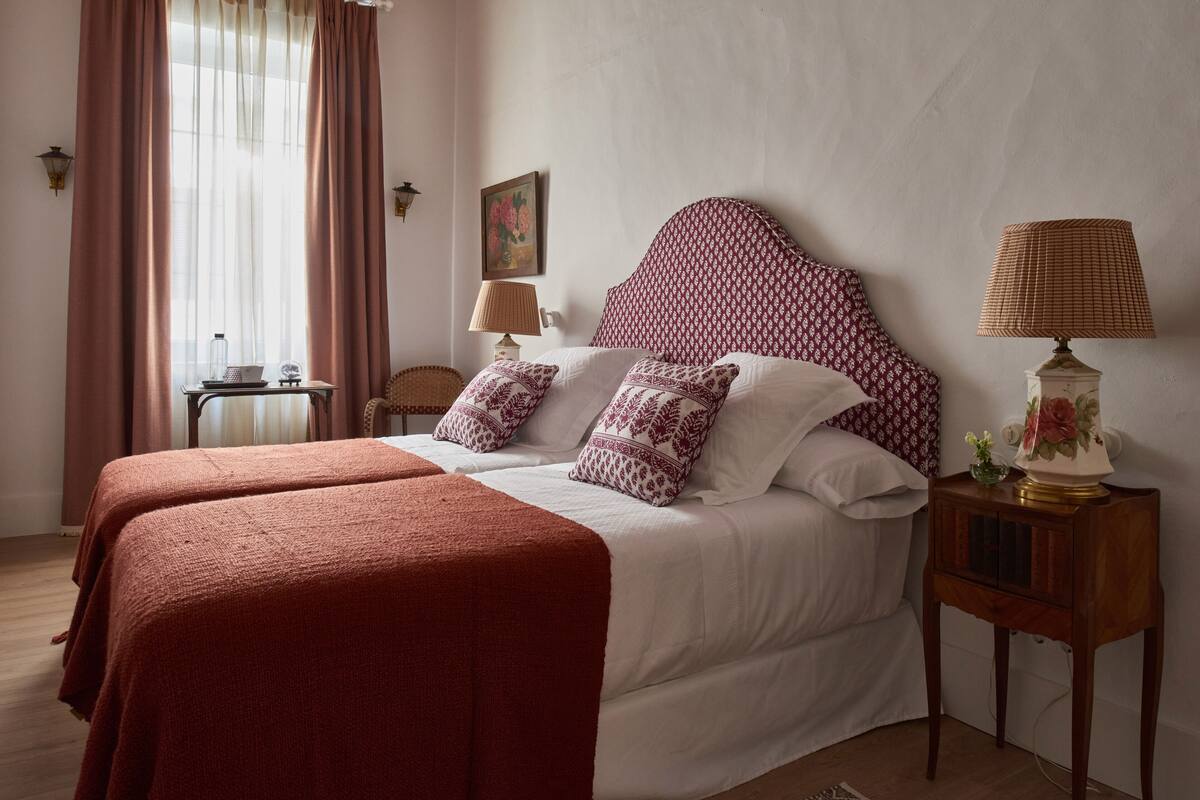 A double bed with a patterned headboard, two bedside tables with lamps, and a window with curtains.