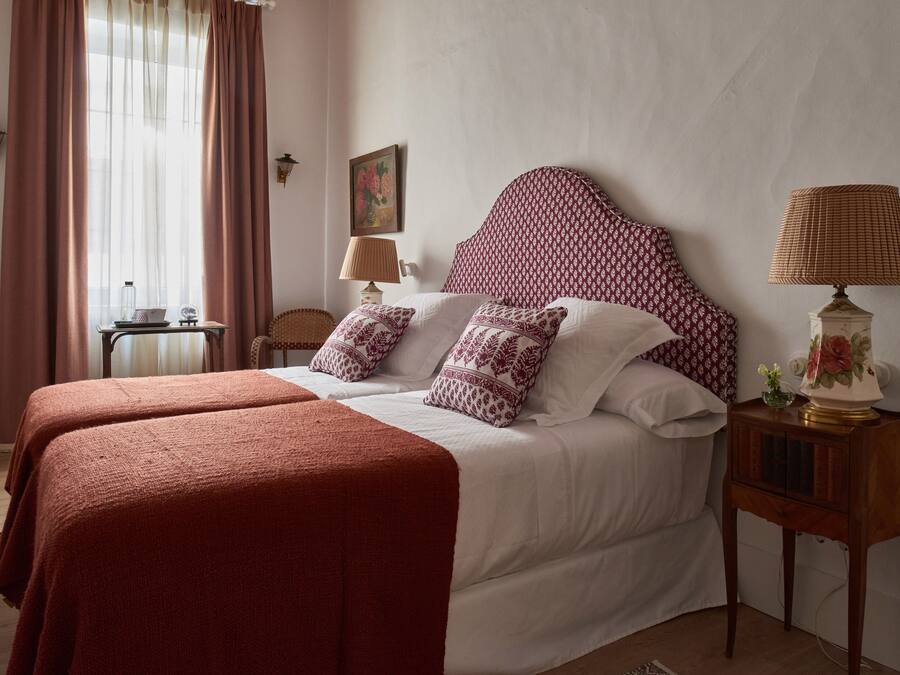 Una cama doble con cabecera estampada, dos mesitas de noche con lámparas y una ventana con cortinas.