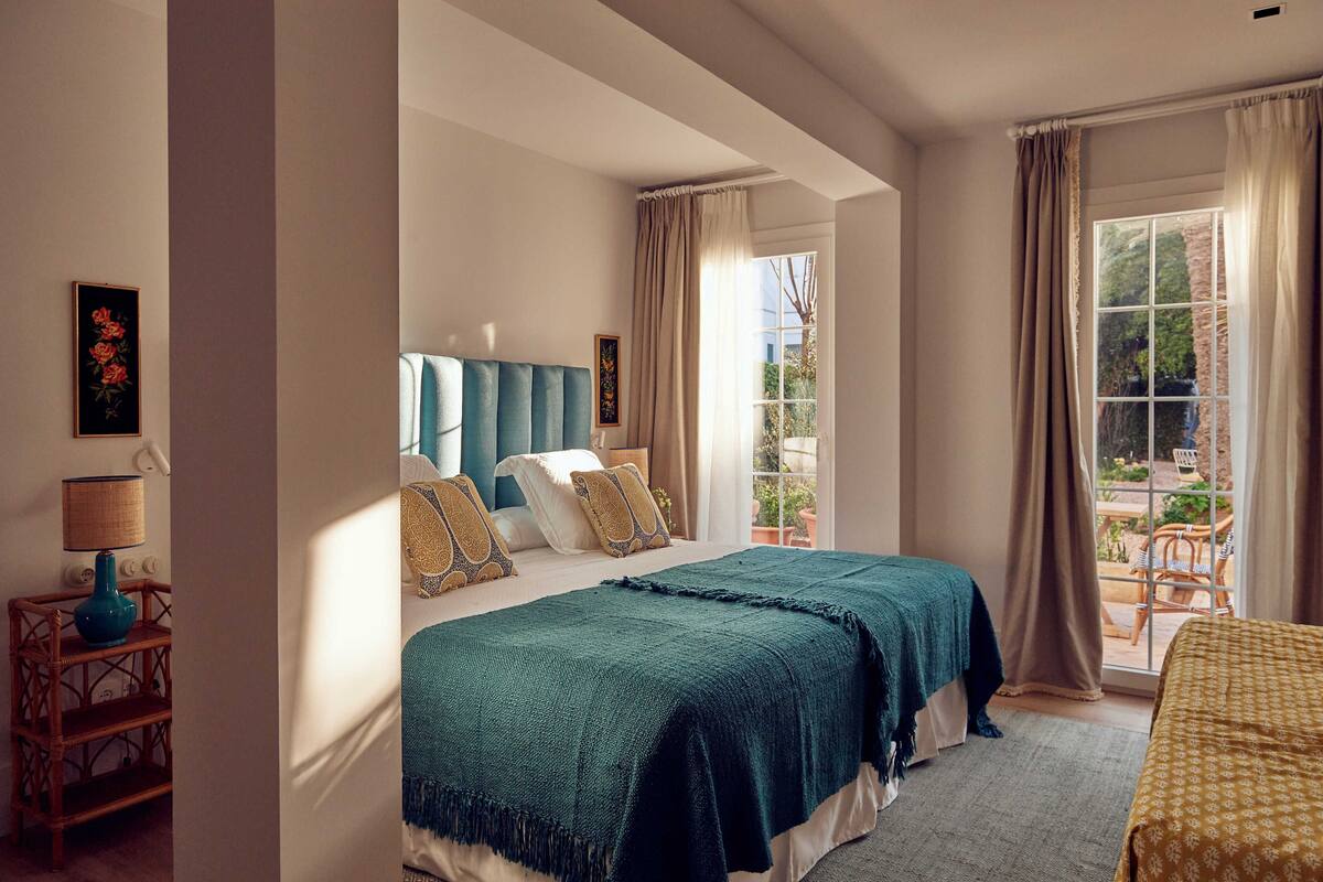 A bedroom with a large bed, a bedside table with a lamp, and a view of the outdoors through a window.