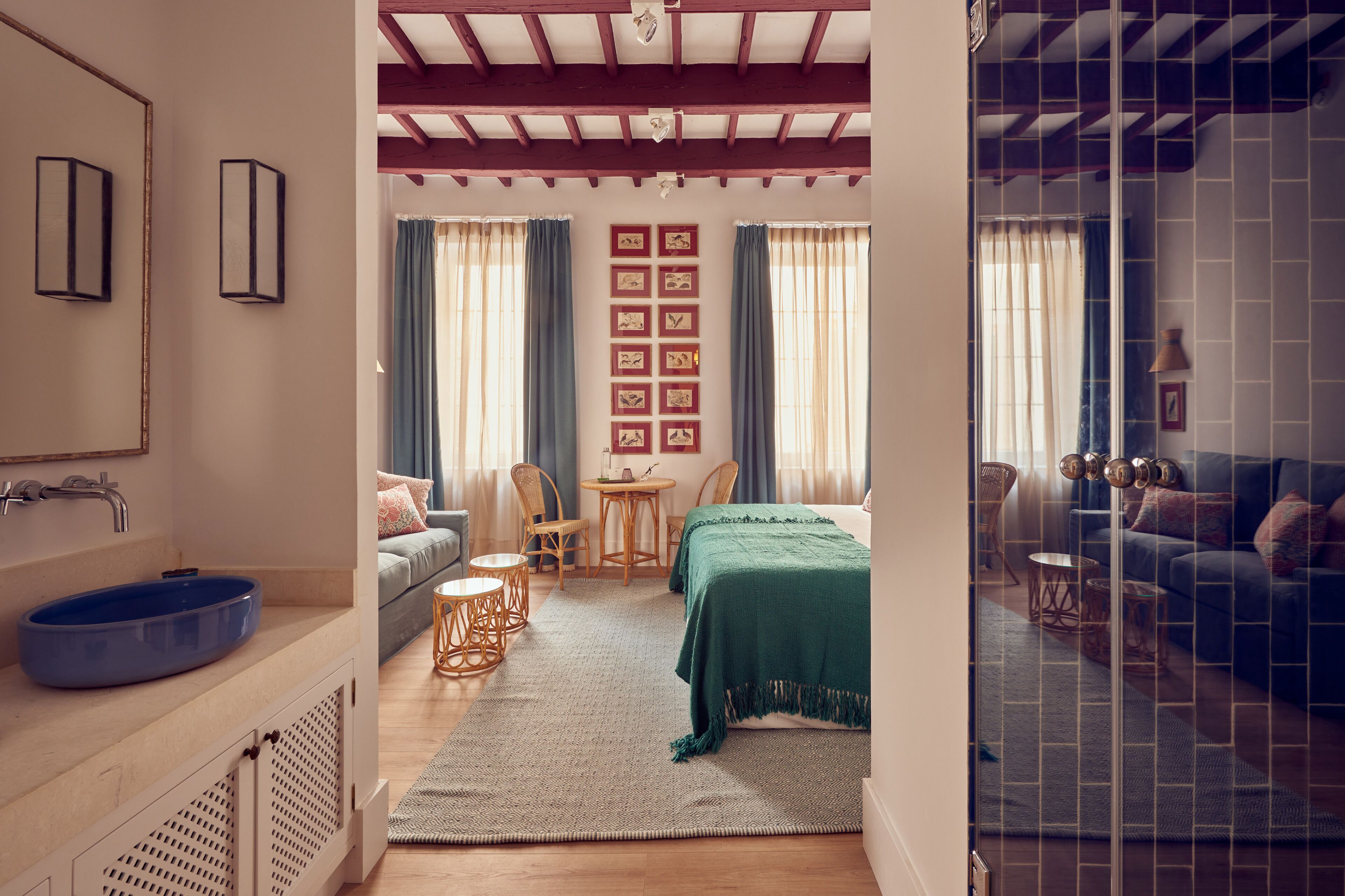 A bedroom with a bed, a small table, two chairs, a sofa, a bathroom with a sink and a blue basin, and a shower area.