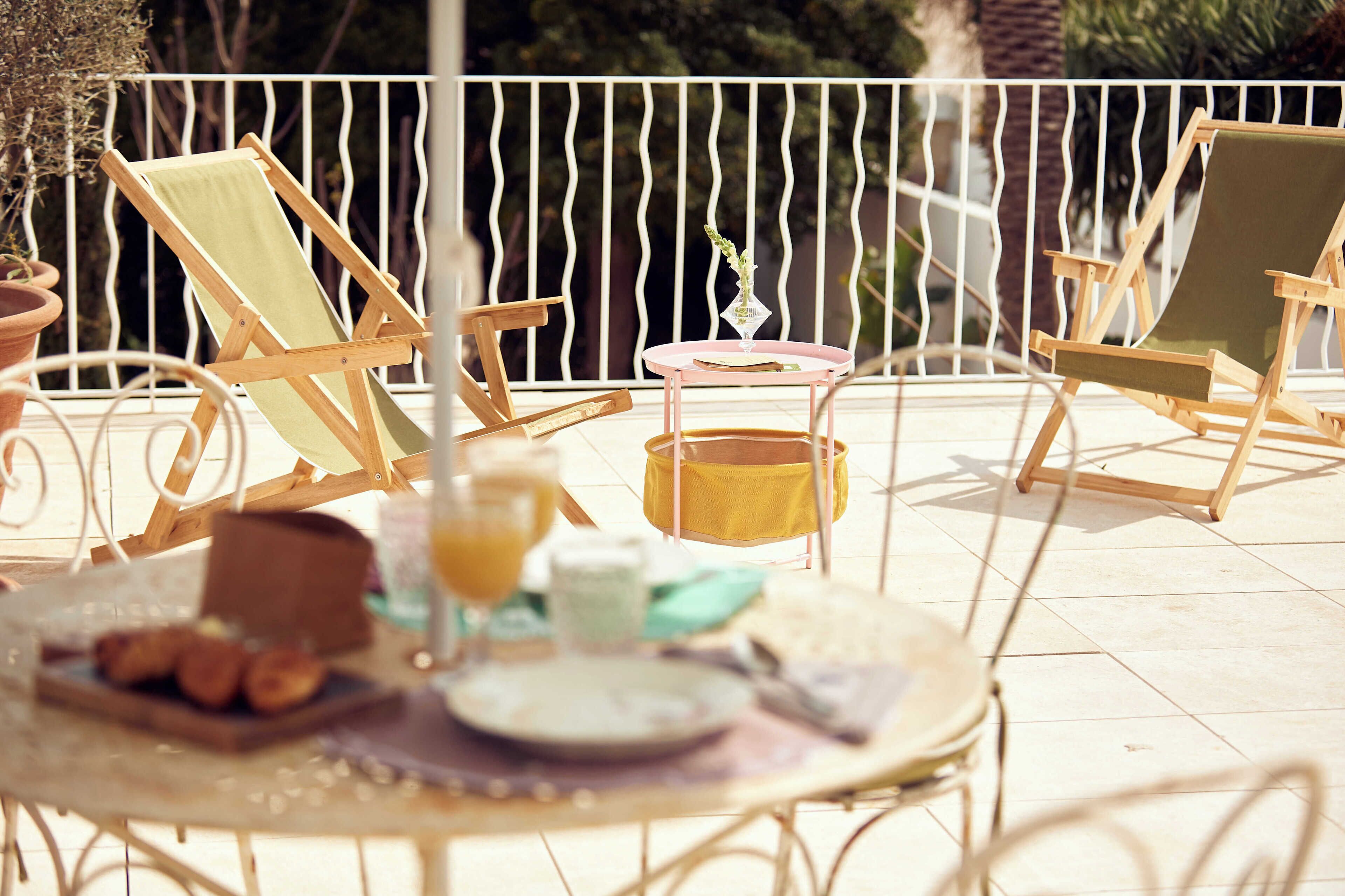 A terrace with two wooden lounge chairs, a small round table with breakfast items, and a potted plant.