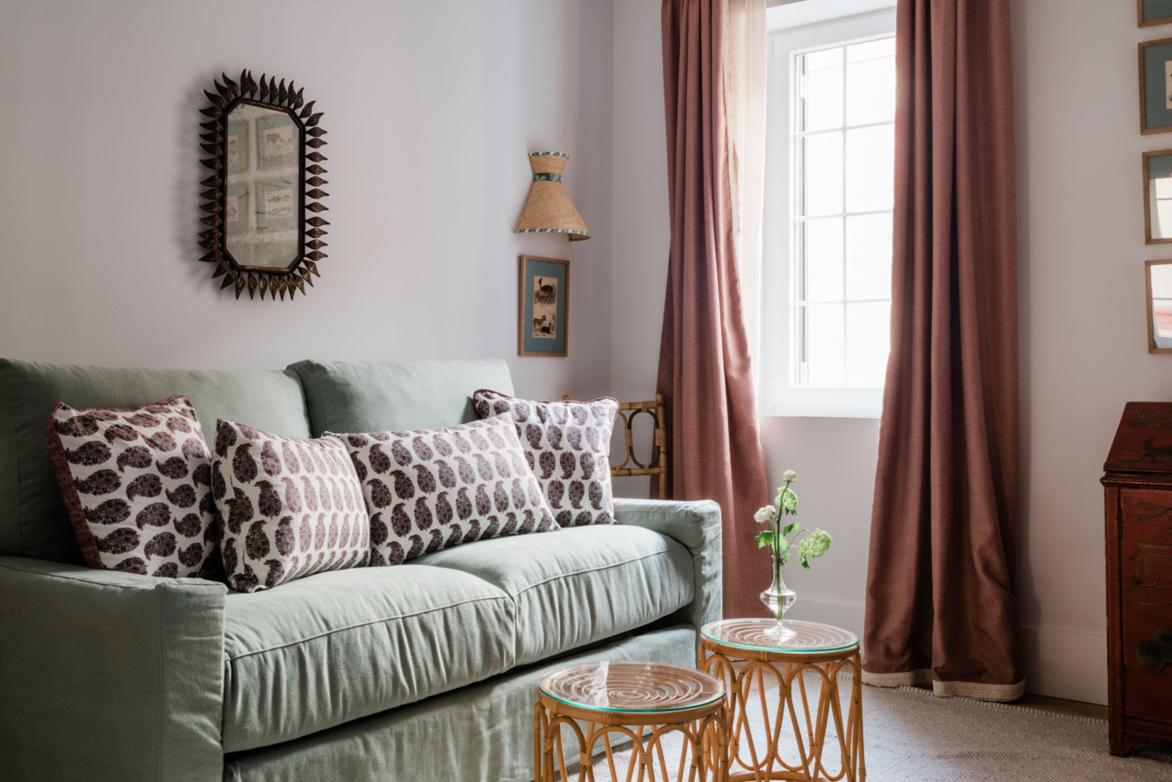 Un salón con un sofá verde, cojines estampados, una pequeña mesa redonda con un jarrón, un espejo con marco de sol y una ventana con cortinas.