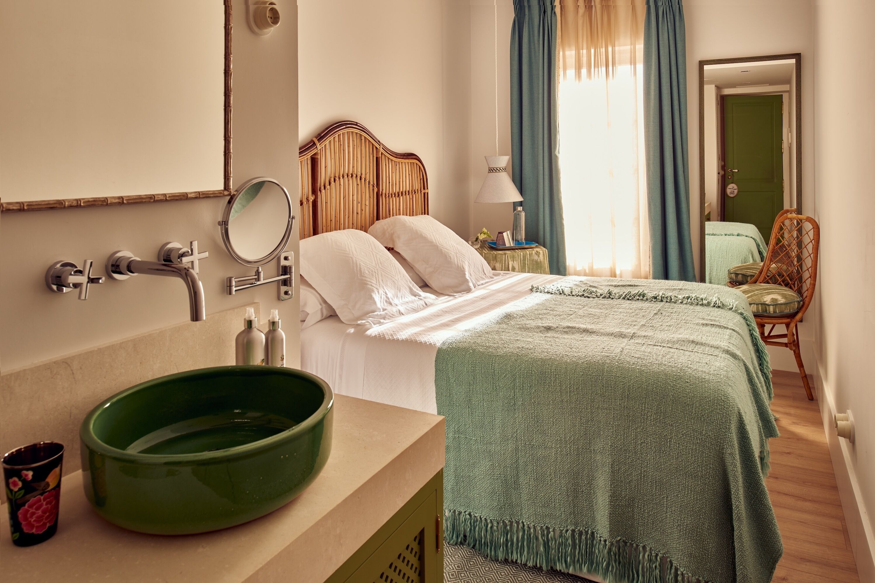 Une chambre d’hôtel avec une tête de lit en bois, un bol vert posé sur la commode, un lit recouvert d’une couverture verte et une chaise près de la fenêtre.