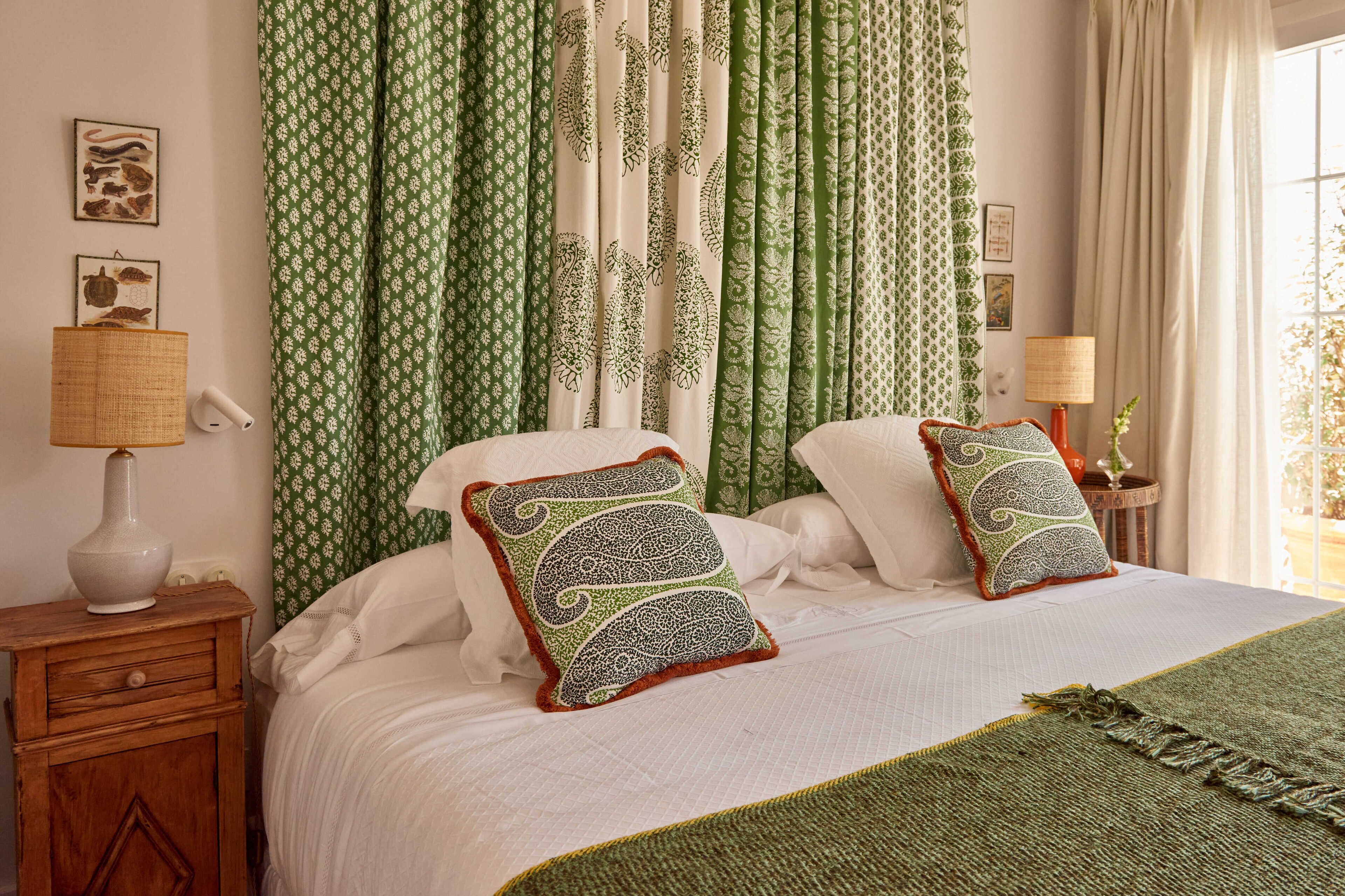 Una cama con dos almohadones decorativos, una lámpara de noche y una cesta tejida sobre una mesita de madera.