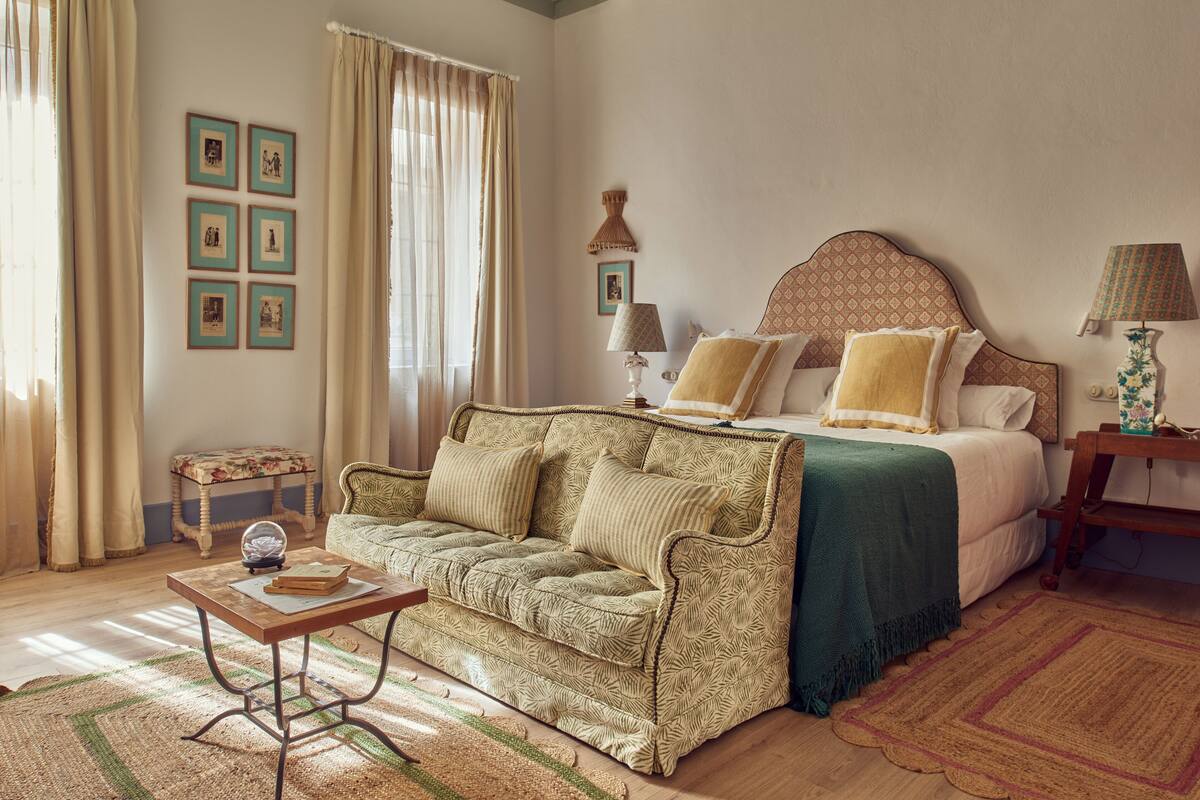A bedroom with a bed, a sofa, a small table, and a lamp.