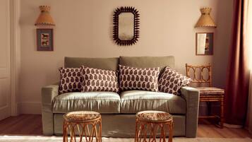 Un salón acogedor con un sofá verde, cojines estampados, dos mesitas de mimbre, una silla de madera y apliques decorativos en la pared.