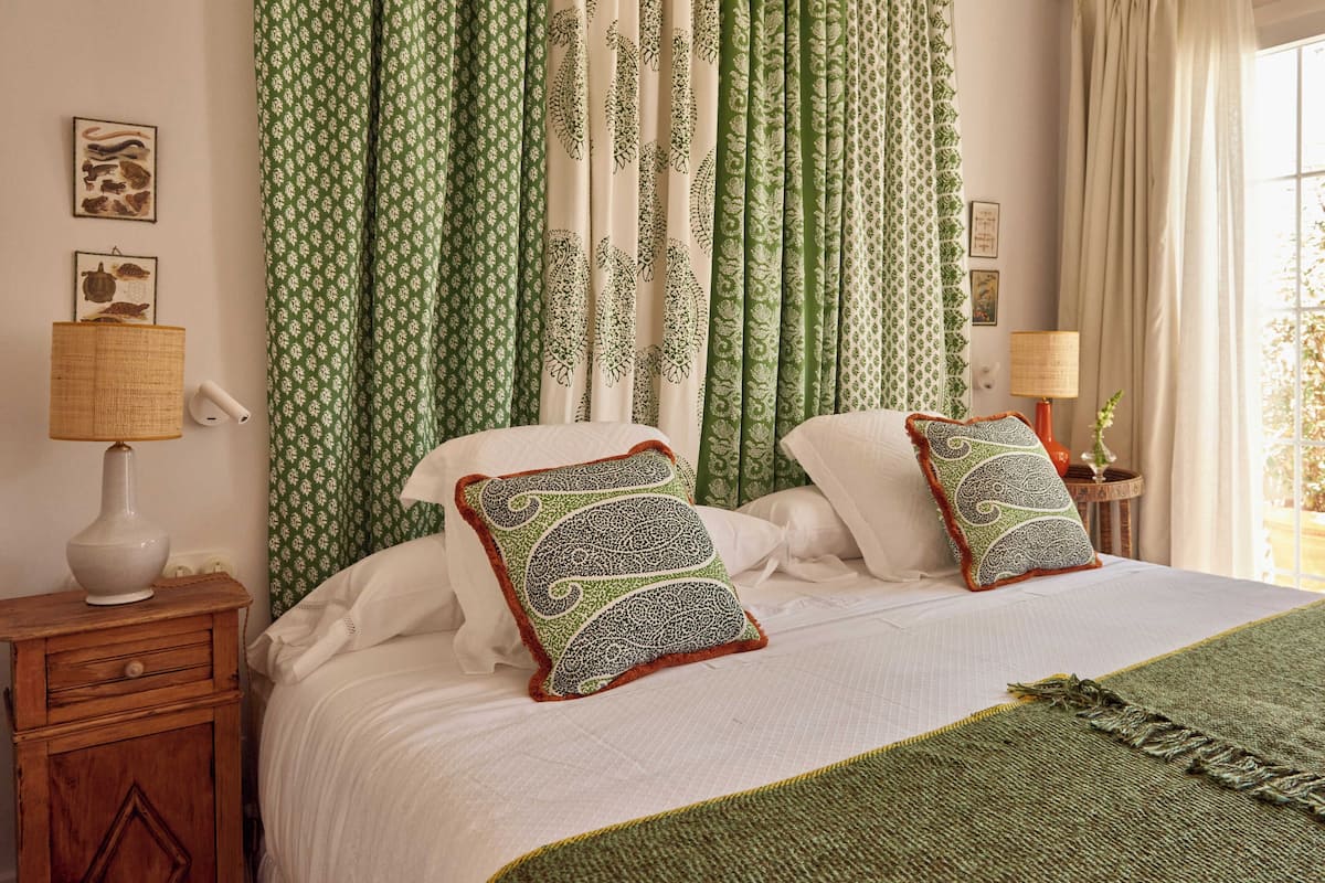 Una cama con dos almohadones decorativos, una lámpara de noche y una cesta tejida sobre una mesita de madera.