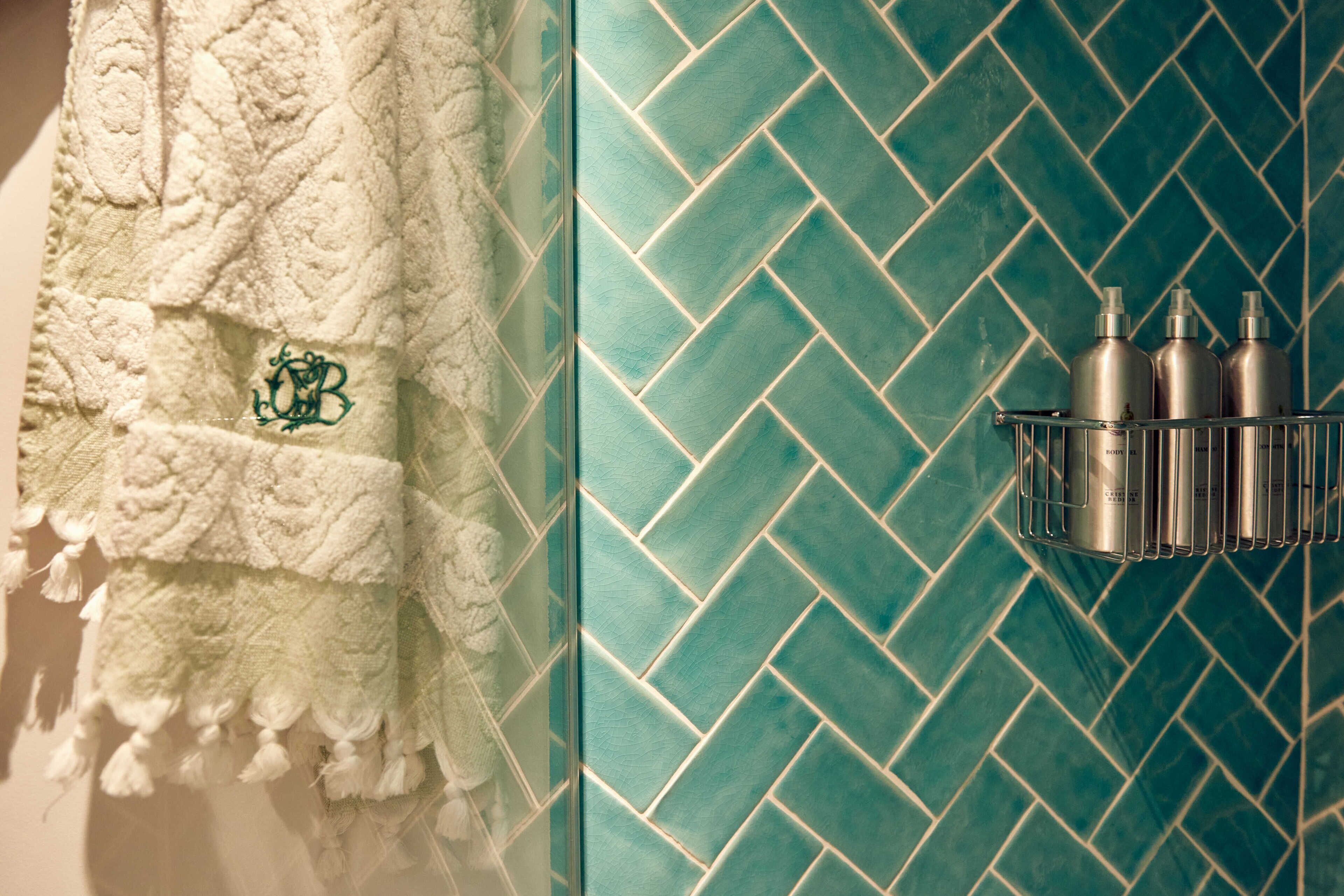 Een badkamer met een witte handdoek aan een rek, een wand betegeld met teal houten visgraatmozaïek en een metalen plank met drie flessen.