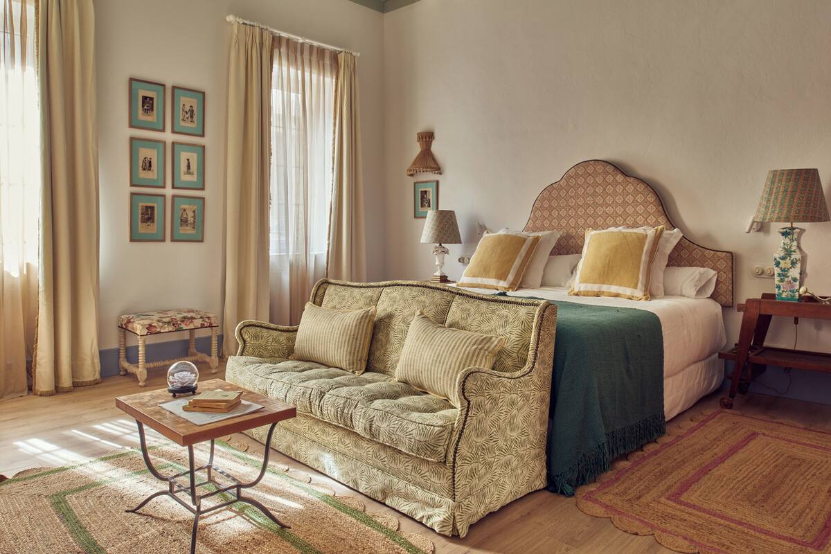 A bedroom with a bed, a sofa, a small table, and a lamp.