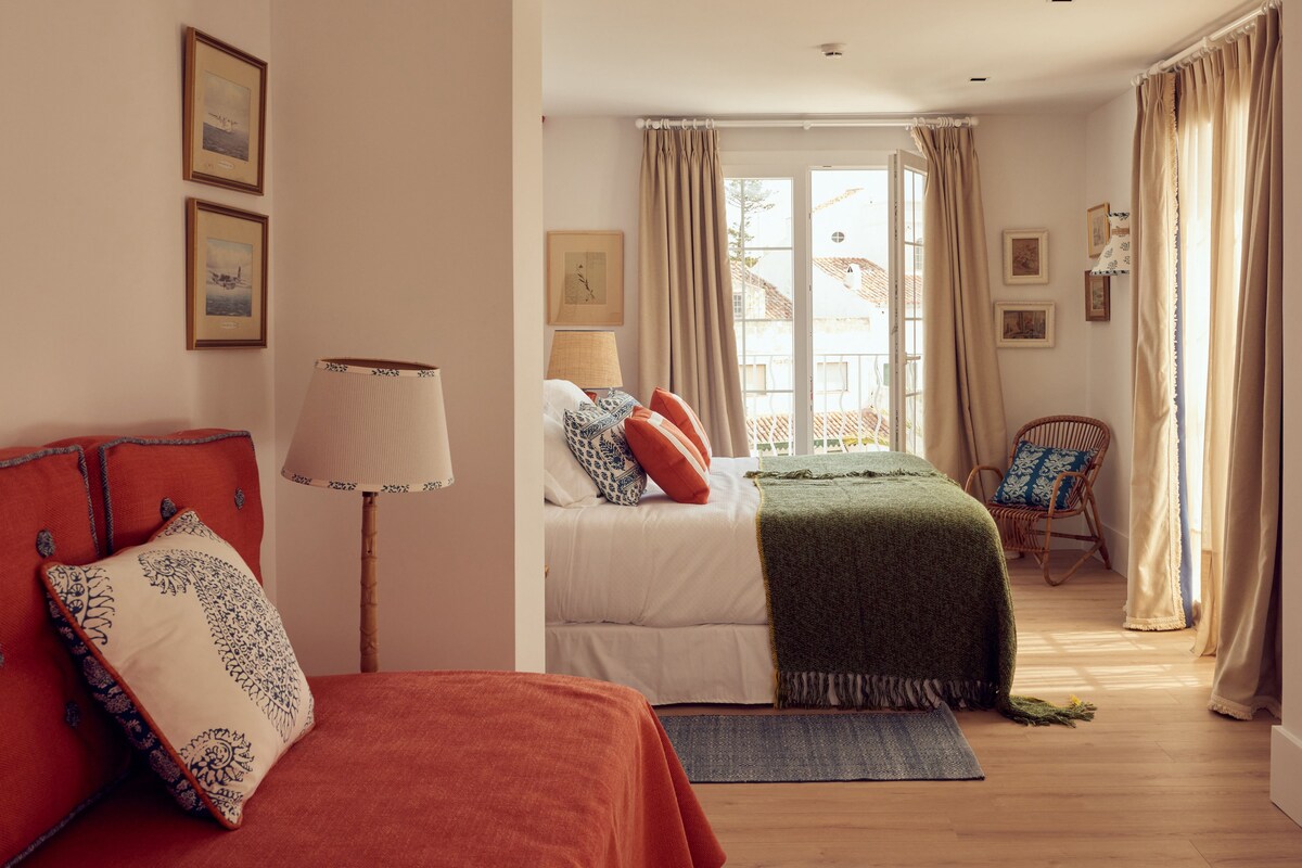 A bedroom with two beds, a chair, a lamp, and a view of the outside.