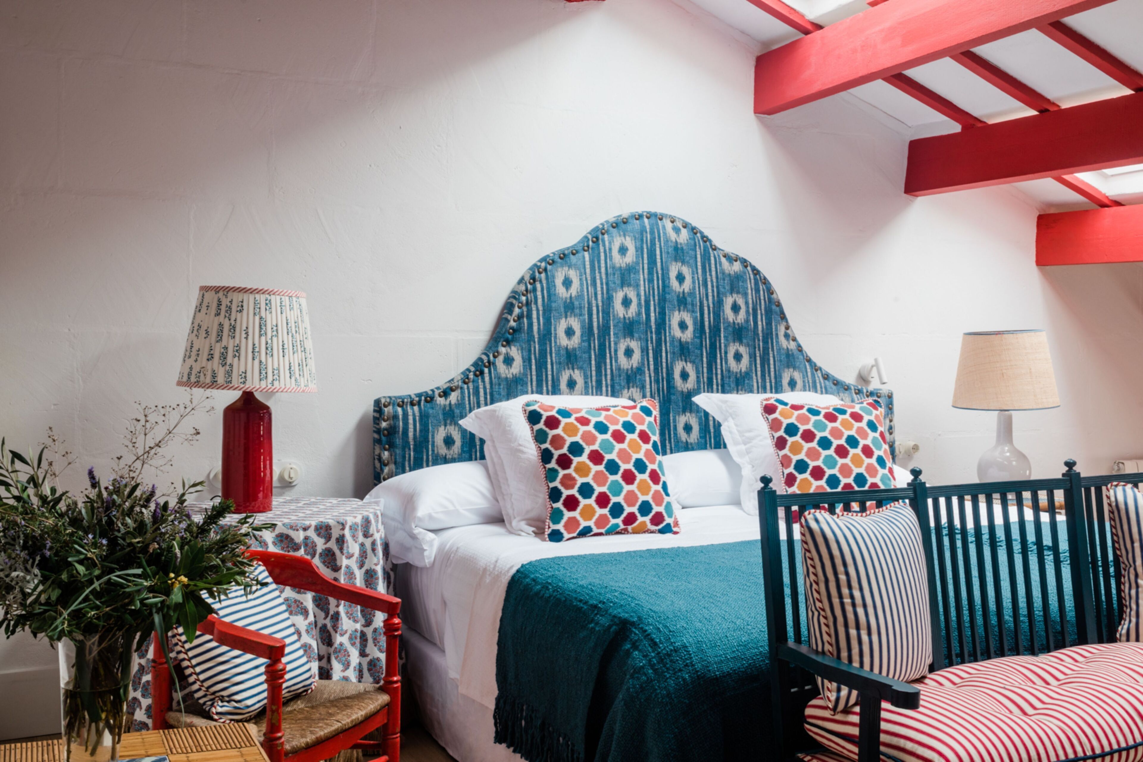 A bedroom with a patterned headboard, a bed with white bedding, two pillows with a polka dot design, a red chair, a small table with a lamp, and a potted plant.