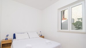 A clean, minimalist bedroom with a single bed, white bedding, a nightstand with a lamp, and a window with a view of a brick building and greenery.