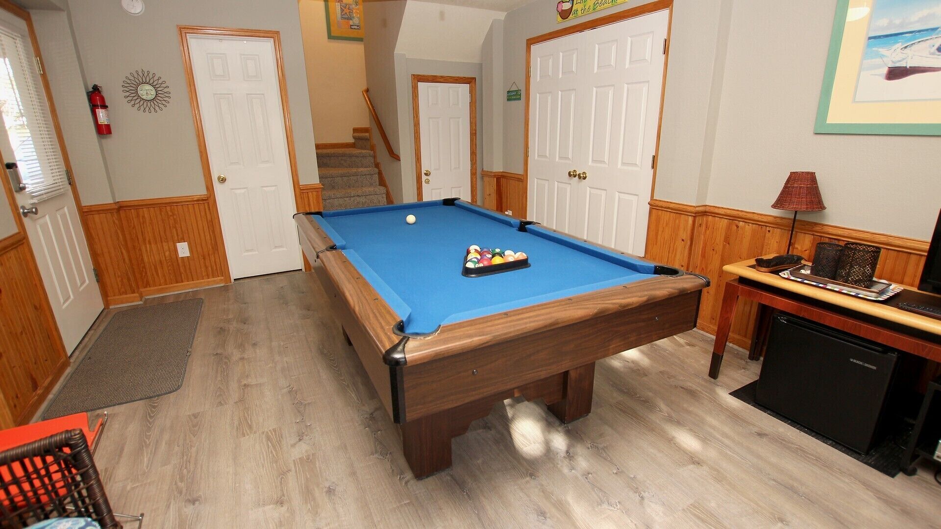 A pool table in a room with wooden floors, a door, a staircase, and a small table with a lamp.