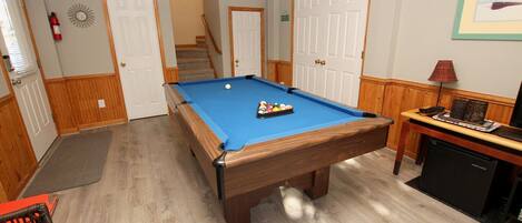 A pool table in a room with wooden floors, a door, a staircase, and a small table with a lamp.