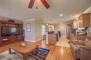 Un vaste espace de vie comprenant un téléviseur, des meubles en bois, un ventilateur de plafond et une cuisine avec des armoires en bois.