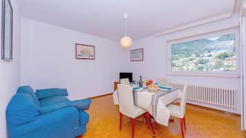 A modern living room with a blue sofa, a dining table with chairs, a hanging light fixture, framed pictures on the wall, and a large window with a view of a town.