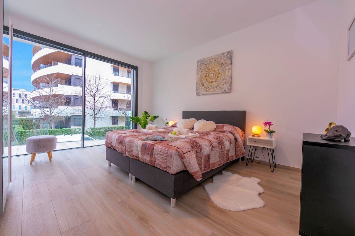 Ein modernes Schlafzimmer mit einem großen Bett, einem kleinen Hocker, einem Nachttisch mit Lampe und Blick auf ein Gebäude durch große Fenster.